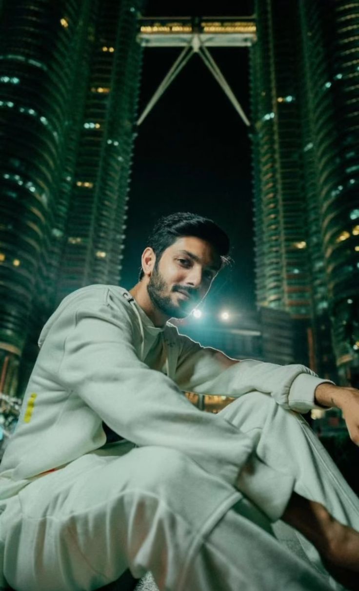 a man sitting on the ground in front of some tall buildings at night with his arms crossed