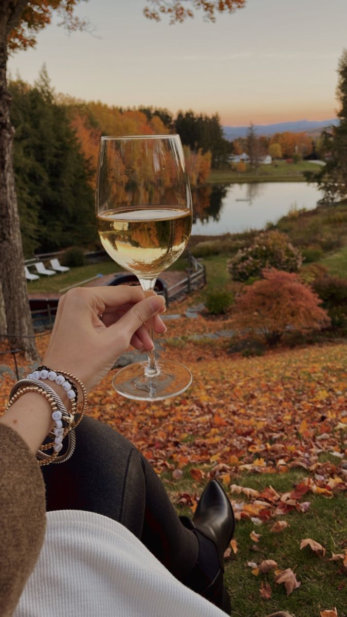 a person holding a wine glass in their hand while sitting on the ground with autumn leaves around them