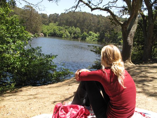 a woman sitting on the ground next to a lake with trees around her and a red bag in front of her