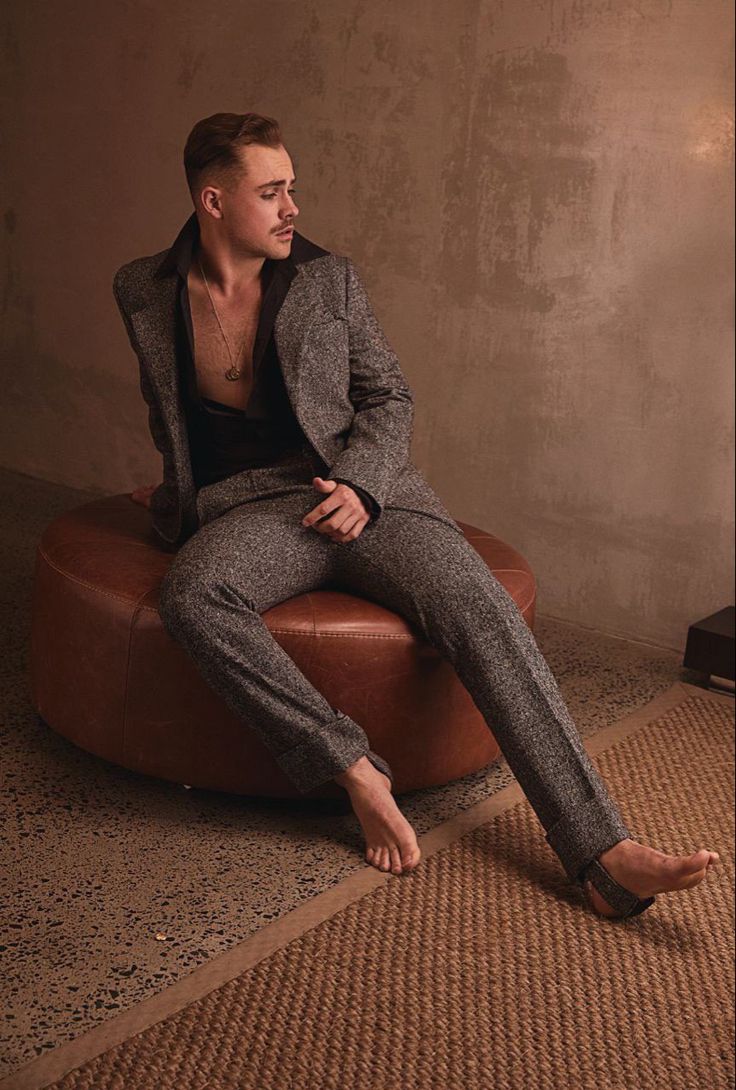 a man sitting on top of a round ottoman in front of a wall and wearing a suit