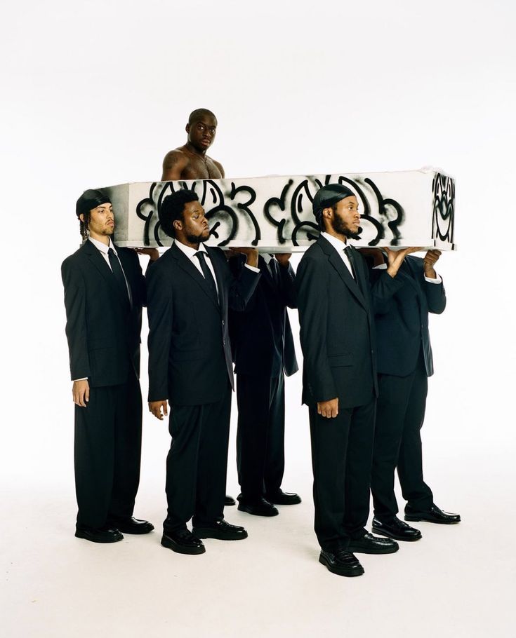 four men in suits are holding up a skateboard with graffiti on it and one man is sitting on top of the board