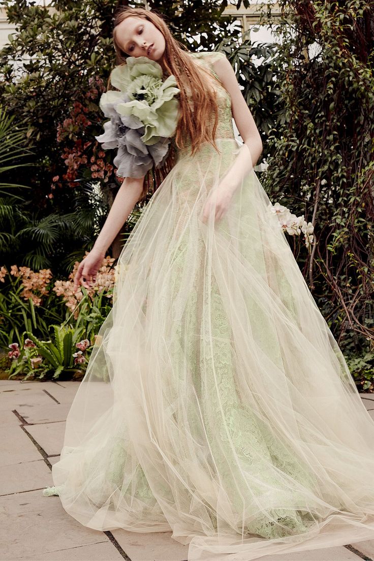 a woman in a green dress is standing near some bushes and flowers with her hands on her hips