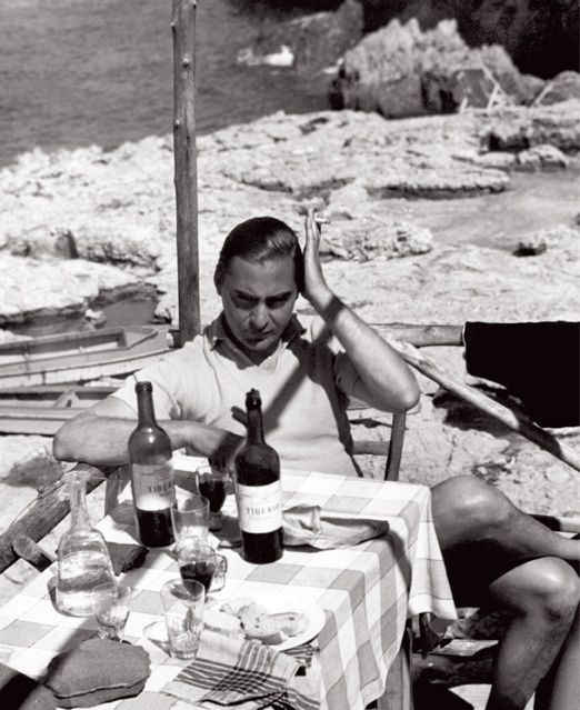 a man sitting at a table with two bottles of wine in front of him on the beach