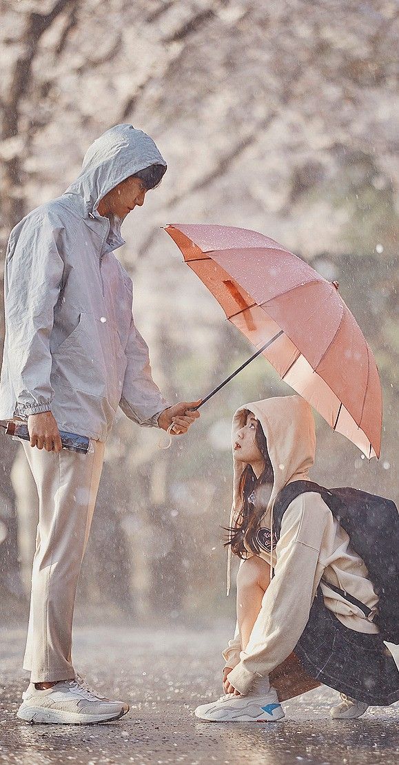 two people standing in the rain with an umbrella over their heads and one person kneeling on the ground