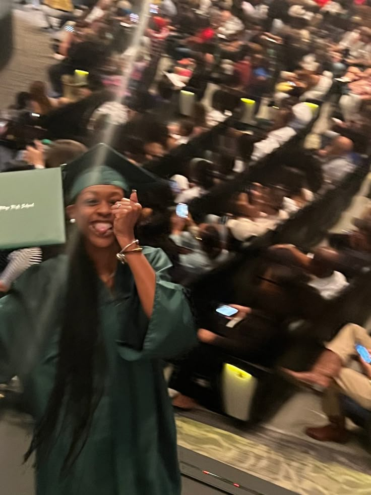 a woman in a graduation cap and gown holding up a laptop computer while standing in front of an auditorium full of people