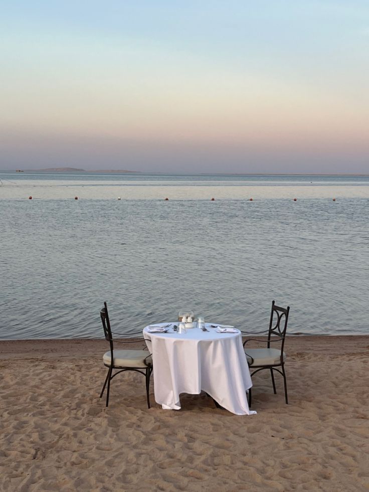 an empty table on the beach with two chairs and a white tablecloth set up