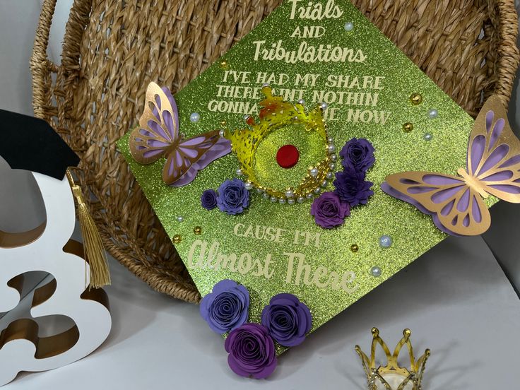 a green graduation cap with purple flowers and butterflies on it sitting next to a basket