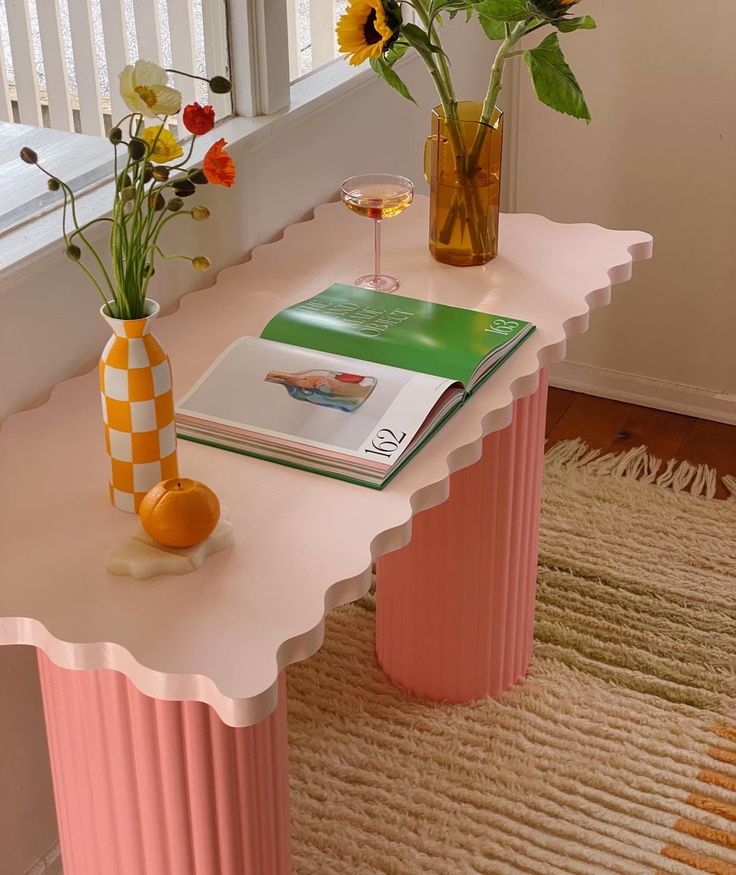 a pink table with flowers and a book on it next to a window sill