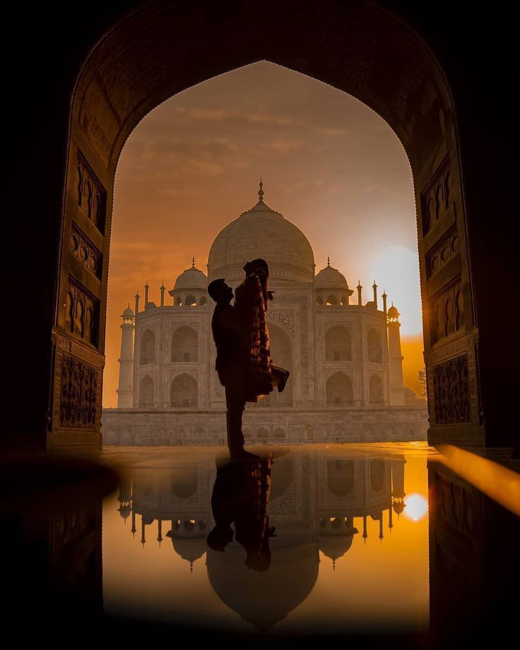 the silhouette of two people standing in front of a building with an arch at sunset