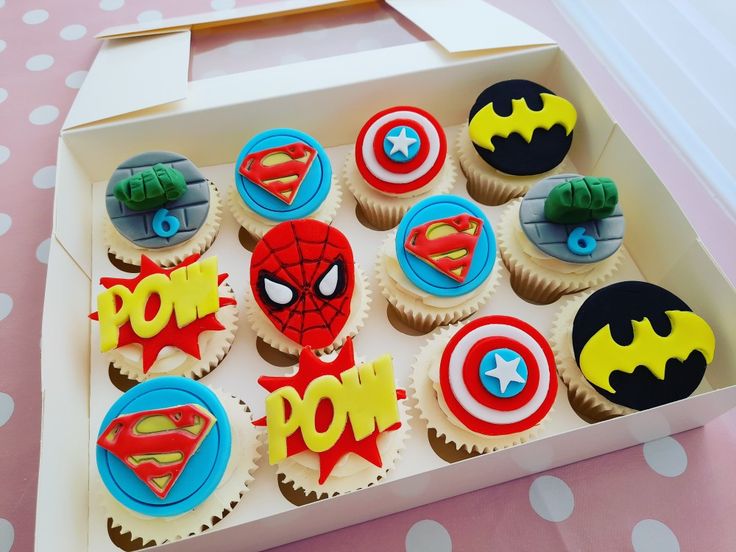 cupcakes decorated like superheros are in a box on a pink tablecloth