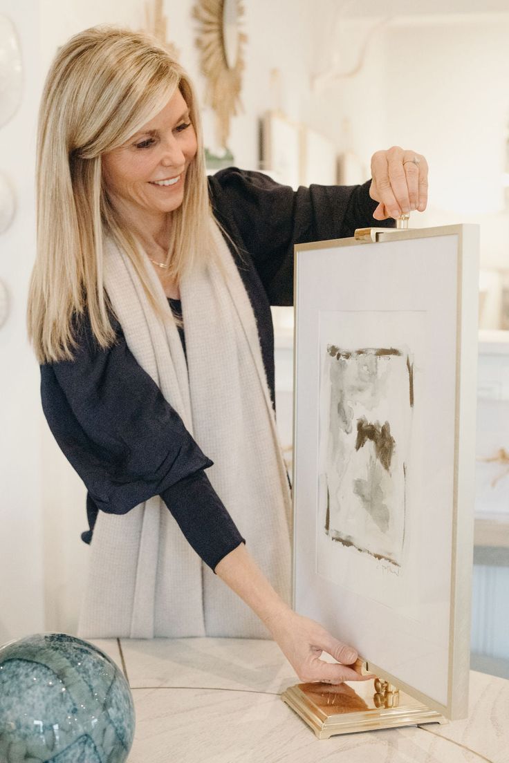 a woman holding up a painting in front of a table