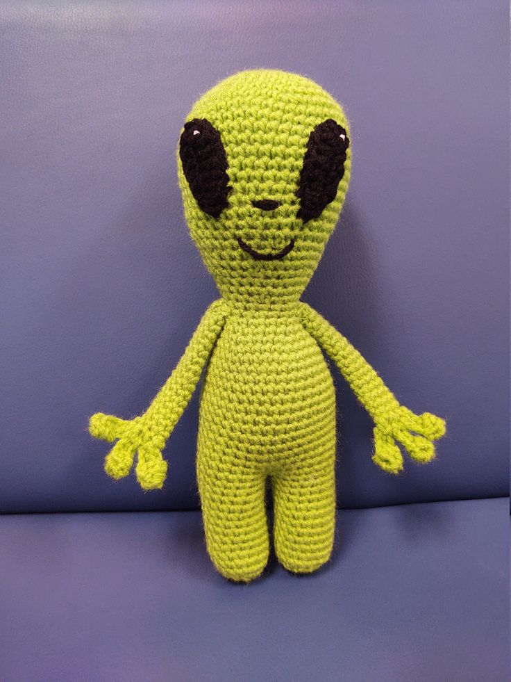 a crocheted alien doll sitting on a purple surface with its eyes wide open