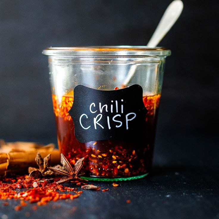 a jar filled with chili and spices next to a wooden spoon on top of a table