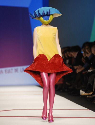 a woman walking down a runway wearing a dress with an unusual hat on her head