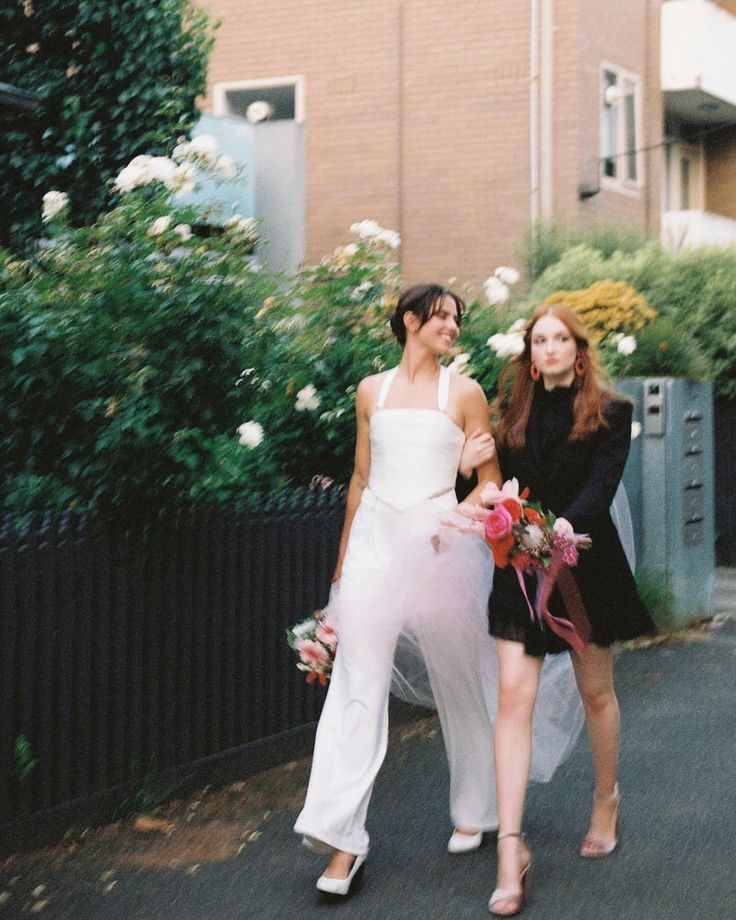 two women are walking down the street with flowers in their hand and one is wearing a white dress