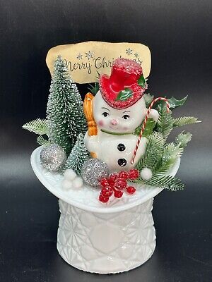 a snowman sitting on top of a white bowl filled with christmas decorations and trees