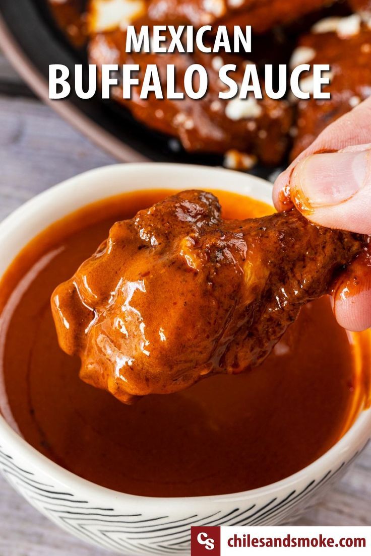 a hand dipping sauce into a white bowl filled with chicken wings in mexican buffalo sauce