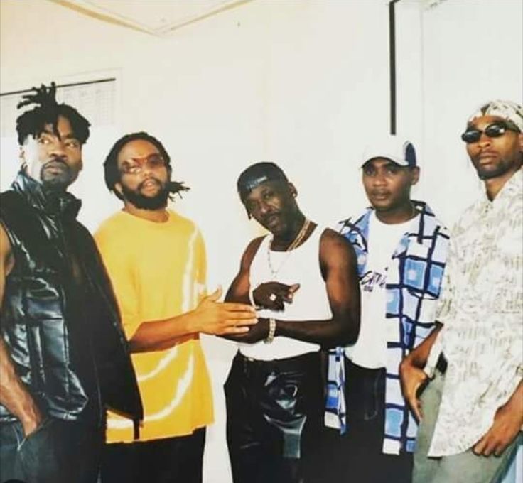 four men are posing for the camera in front of a white wall and one man is pointing at something