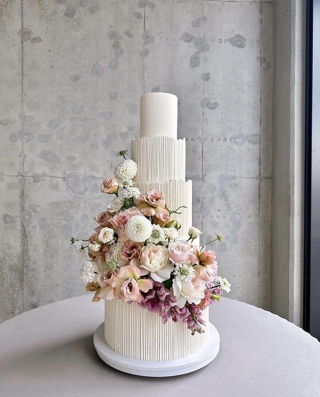 a three tiered white cake with pink and white flowers on the top, sitting on a round table in front of a concrete wall