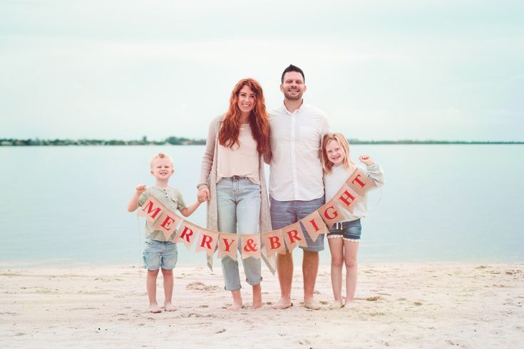 a family standing on the beach holding a banner that says merry and bright