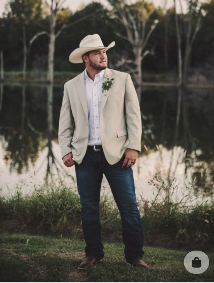 a man wearing a cowboy hat standing in front of a body of water and trees