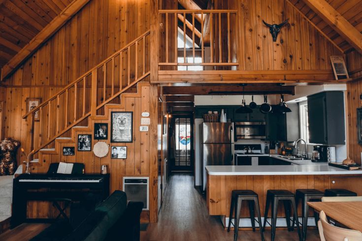 a kitchen and living room in a cabin with wood paneling on the walls, wooden stairs leading up to an upper floor