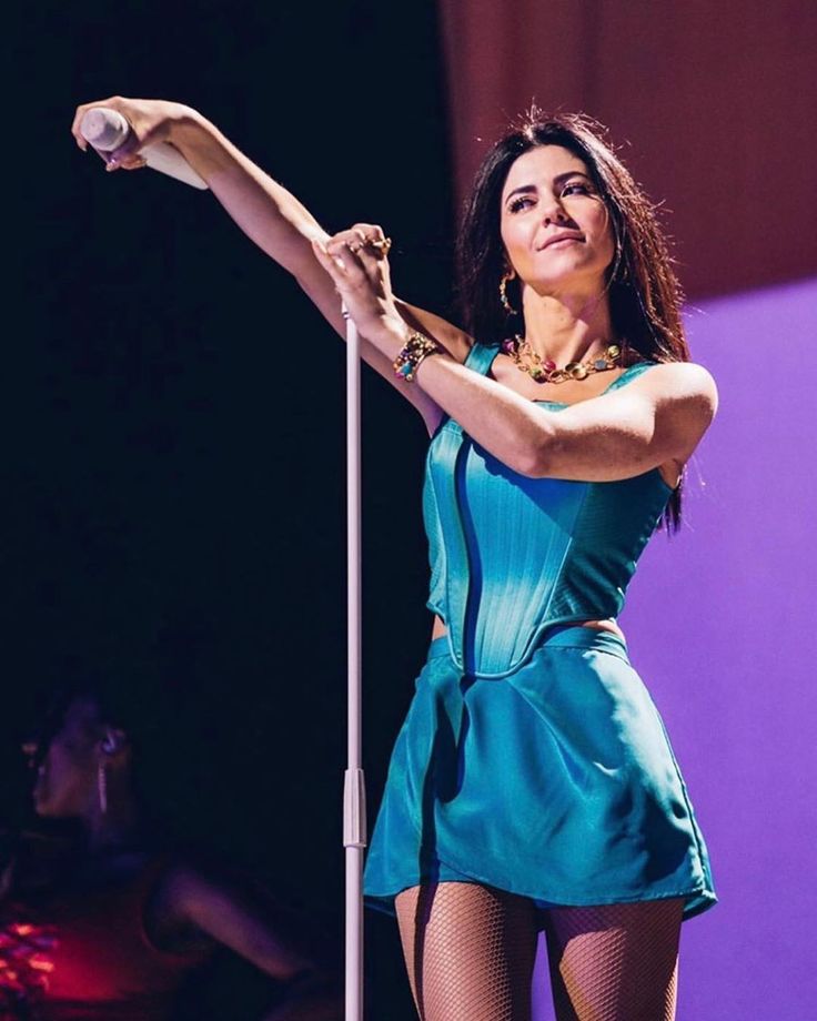 a woman in a blue dress is holding a microphone and standing on stage with her arms outstretched