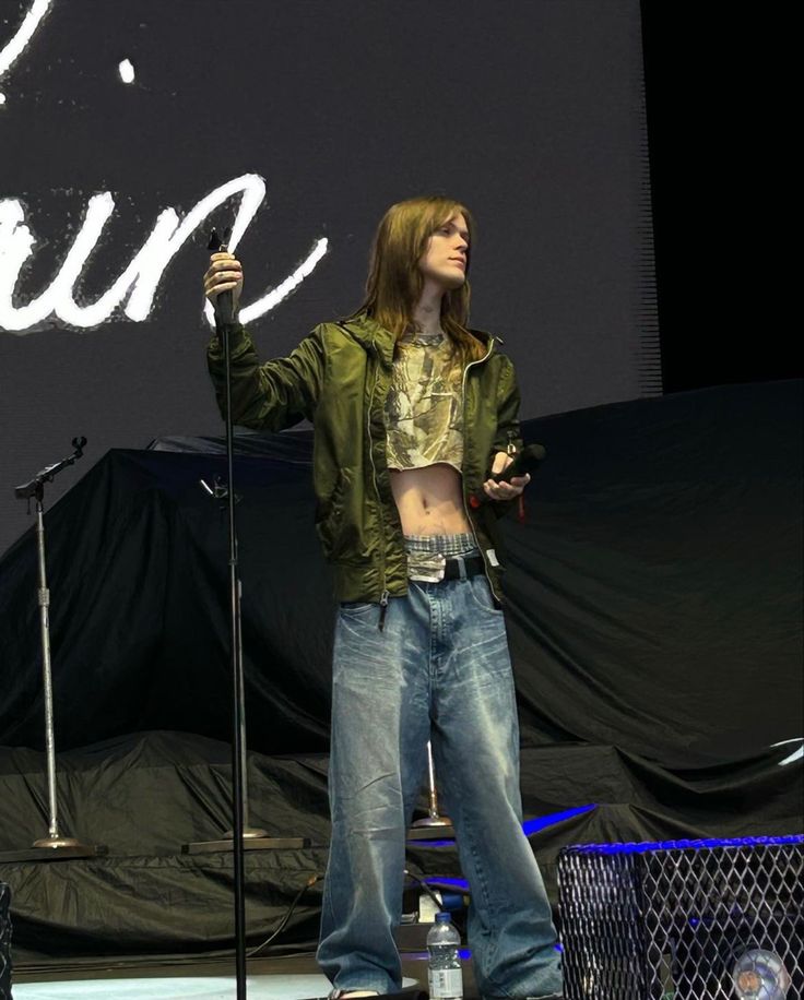 a man standing on top of a stage holding a microphone in his right hand and wearing jeans