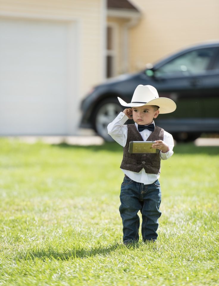 a young boy wearing a cowboy hat and vest standing in the grass with a book