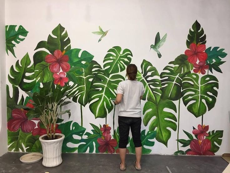 a woman standing in front of a wall painting with tropical leaves and flowers on it