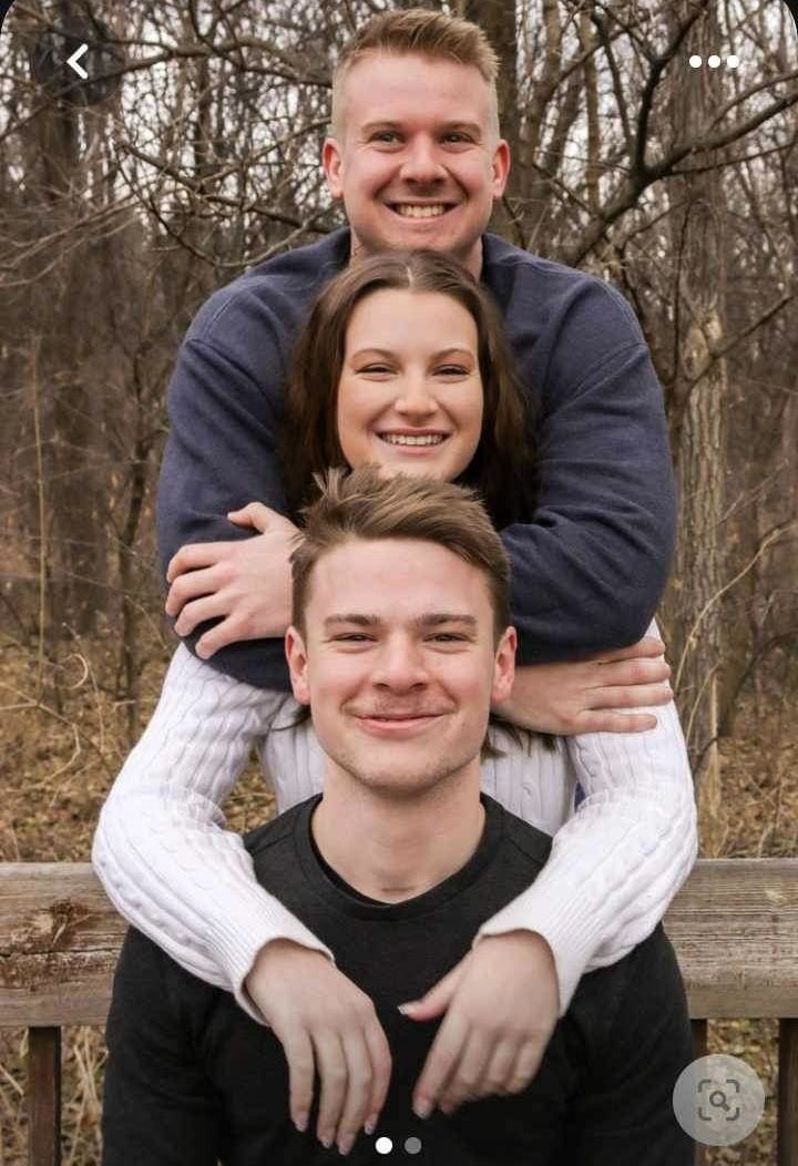 three people sitting on top of each other in front of trees and bushes, with one man holding the woman's shoulders