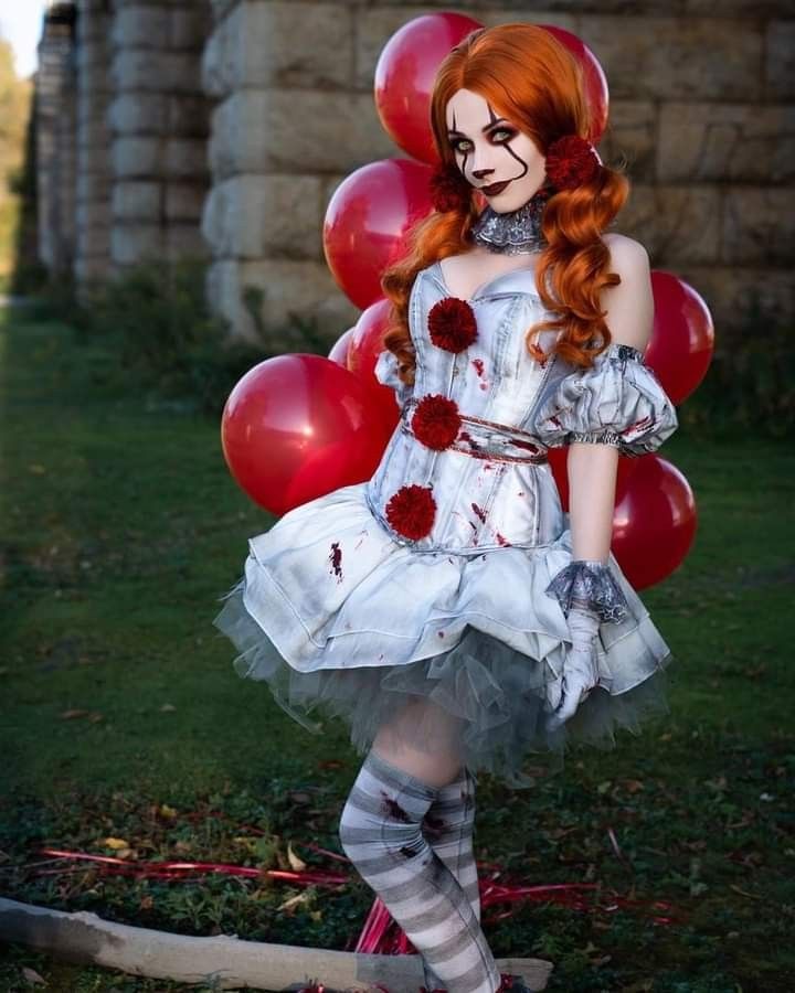 a woman dressed up as a clown with red hair and makeup, standing in front of balloons