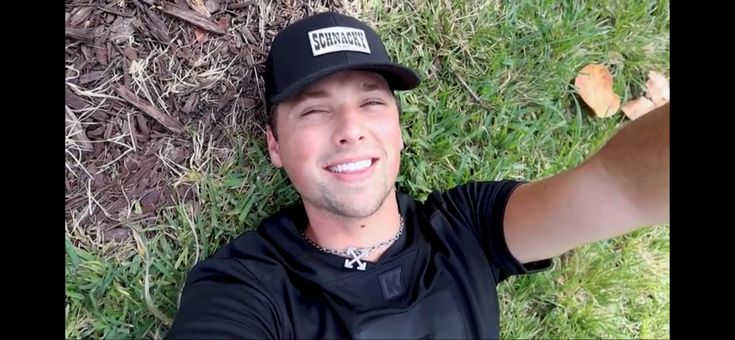 a man laying in the grass wearing a hat and holding his arm out to take a selfie