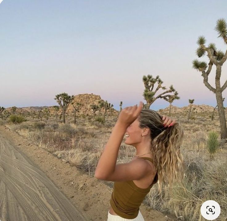 a woman standing in the middle of a dirt road next to a desert landscape with cacti