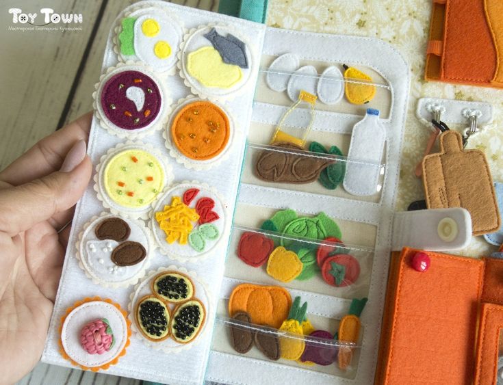 a person is holding an open book with felt food on it and sewing supplies in the background