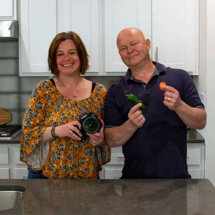 two people standing in a kitchen holding carrots and broccoli stalks with one person holding a camera