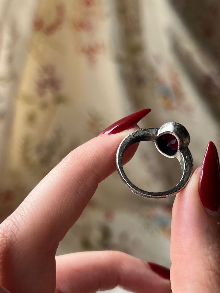 A gorgeous and vampy adornment that is a custom Curio original! These hand-made pieces are a true treasure, for they are one of a kind and crafted with intention to marry the aesthetics of the past, with the modern stylings of the present. This hammered silver garnet ring features a stunning blood-red garnet cabochon set in striking sterling silver. The band is simple and understated, with fine scrolling foliate detail that bring a gentle glamour to the silhouette. There is versatility in this design as well – pair it with a casual or formal look, or even go another step ahead and loop it through a delicate chain as a charm. Either way, this Curio Original is a magnificent addition for any jewelry lover’s collection! Materials and Features: Sterling Silver, Garnet Cabochon Age/Origin: Curi Spiritual Open Ring Jewelry With Ring Detail, Artisan Open Ring Gemstone Jewelry, Unique Ruby Gemstone Jewelry, Bohemian Promise Ring Jewelry, Hand Forged Fine Jewelry Promise Ring, Hand Forged Vintage Jewelry, Hand Forged Vintage Promise Jewelry, Hand Forged Open Ring For Promise, Elegant Handmade Metal Crystal Ring