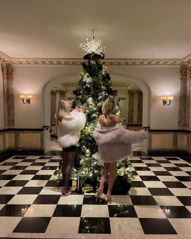 two women standing in front of a christmas tree with lights on the top and black and white checkered floor