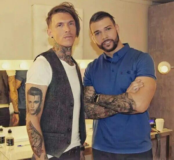 two men standing next to each other with tattoos on their arms and shoulder, both looking at the camera