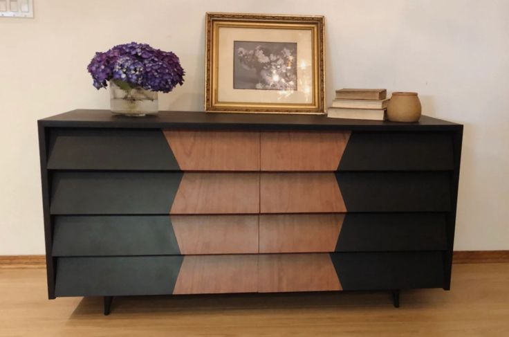 a black and brown dresser with purple flowers on top next to a framed photo in a gold frame