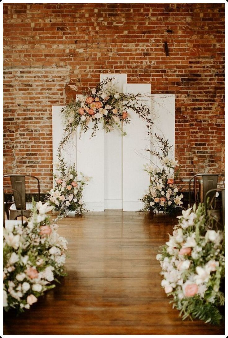 an indoor ceremony with flowers and greenery on the aisle, set up in front of a brick wall