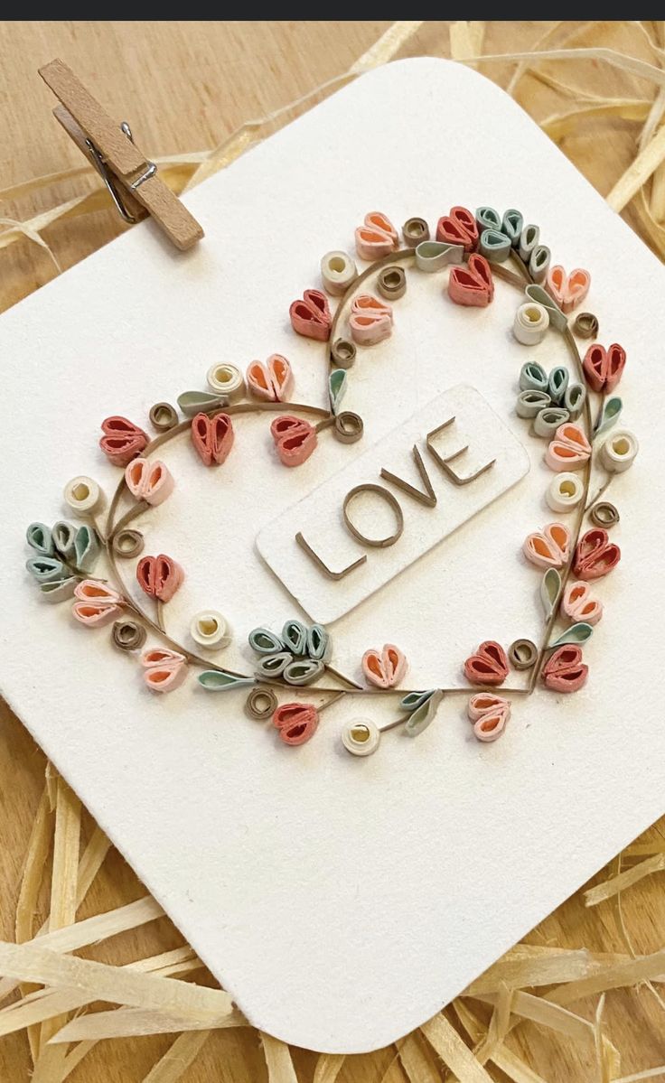 a heart shaped card with the word love surrounded by flowers and leaves, on top of straw