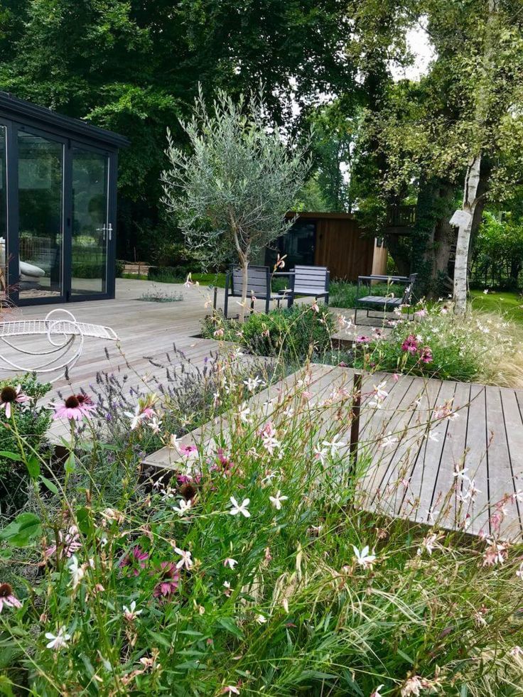 a wooden deck surrounded by lots of flowers and greenery next to a glass walled building