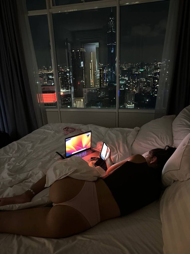 a woman laying in bed with a laptop on her lap and looking out the window