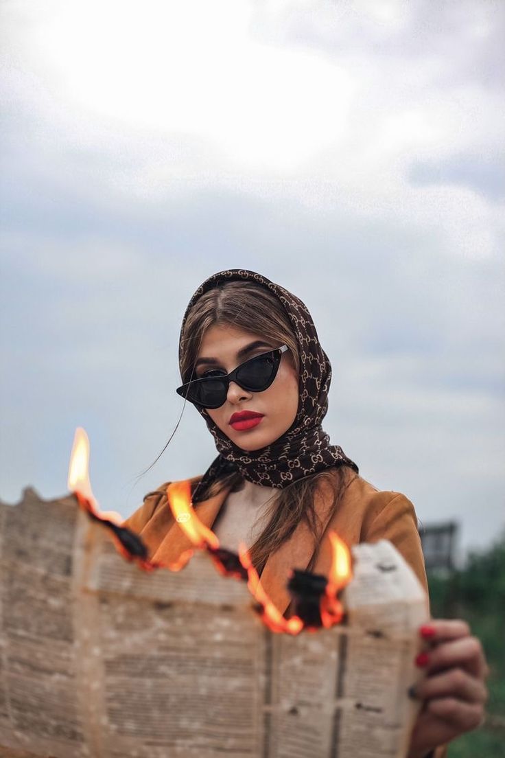 a woman wearing sunglasses and scarf holding a newspaper with flames coming out of the pages