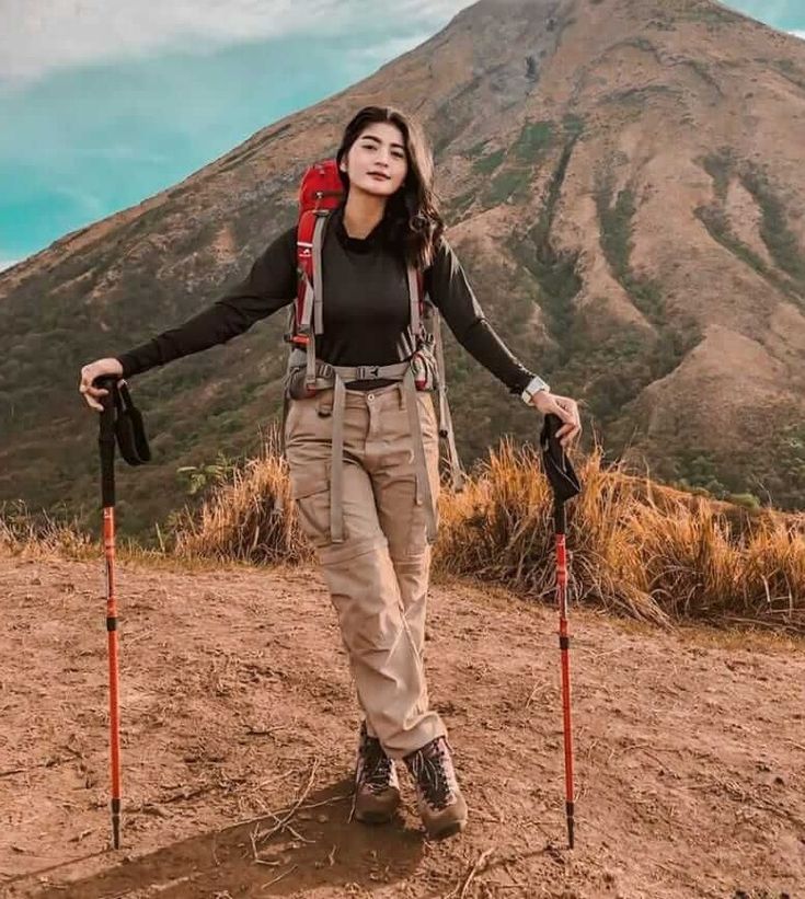 a woman standing on top of a dirt hill holding two poles and wearing a backpack