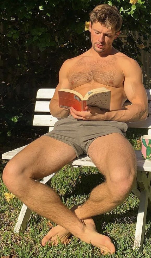 a shirtless man sitting on a bench reading a book in the grass with his legs crossed