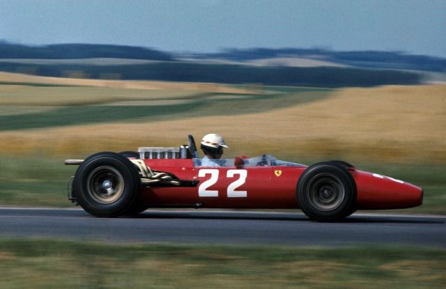 a man driving a red race car down a road in front of some hills and grass