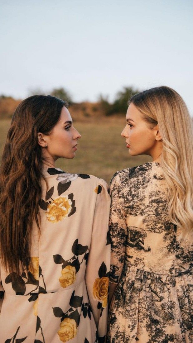 two young women standing next to each other in front of a field with grass and trees