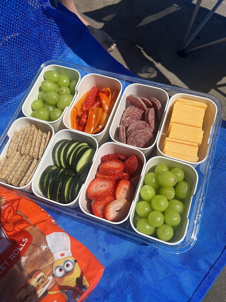 an assortment of fruits and cheeses in plastic containers on a blue table cloth next to a bag of crackers